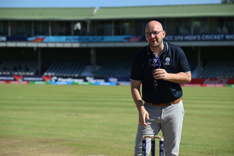 Andrew Leonard holding a microphone while presenting at a cricket ground