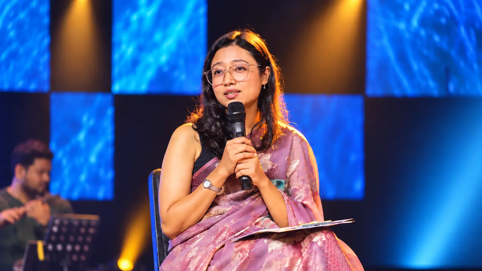 Munu Adhikari speaking into a microphone at a stage event, wearing a lavender sari
