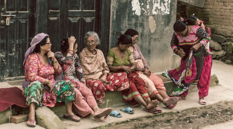 Older Nepali women sitting together near a home, representing ageing parents and community support