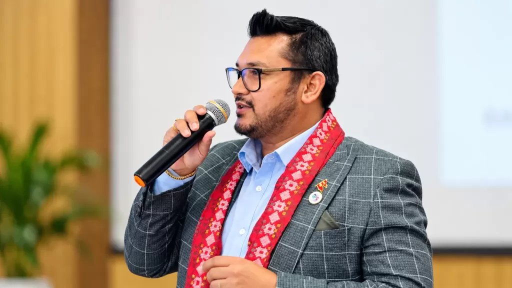 Ramesh Shrestha speaking into a handheld microphone at a community event, wearing a grey checkered blazer and a red traditional scarf